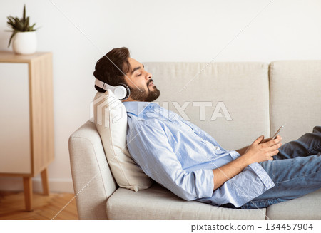 Young Arab Man In Wireless Headphones Relaxing On Couch With Smartphone, Calm Eastern Man Listening Favorite Music Or Audiobook While Resting On Sofa In Living Room, Side View With Free Space 134457904