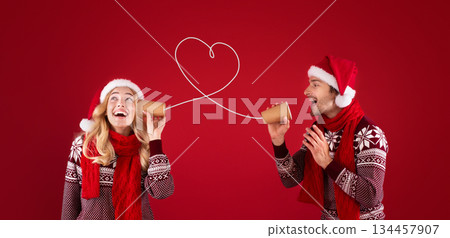 Christmas love message. Cute couple in Santa hats and sweaters talking on tin phone over red background, panorama 134457907