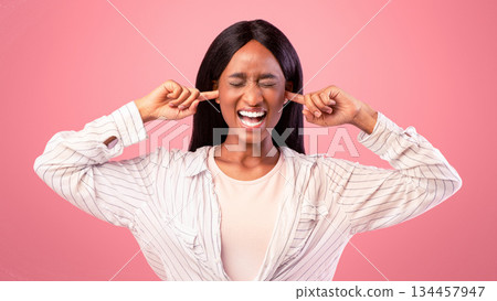Portrait of irritated young black woman closing ears with fingers and shutting her eyes on pink studio background. African American lady angry over loud noise, having headache or suffering from stress Portrait of irritated young black woman closing ears with fingers and shutting her eyes on pink studio background. African American lady angry over loud noise, having headache or suffering from stress 134457947