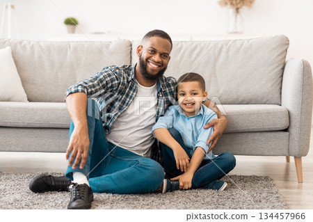 Happy African American Father And Son Embracing Sitting On Floor Posing Smiling To Camera At Home. Portrait Of Young Dad And His Child. Family Bond, Happiness Concept Happy African American Father And Son Embracing Sitting On Floor Posing Smiling To Camera At Home. Portrait Of Young Dad And His Child. Family Bond, Happiness Concept 134457966