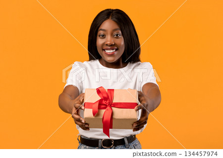 Cheerful young black lady outstretching present towards camera, orange studio background, copy space. Happy african american woman in white t-shirt giving gift box, greeting with birthday Cheerful young black lady outstretching present towards camera, orange studio background, copy space. Happy african american woman in white t-shirt giving gift box, greeting with birthday 134457974