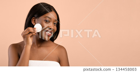 Cheerful black woman holding cotton pad for face cleaning over beige studio background, free space. Happy naked lady cleansing face with toner after shower, enjoying fresh mouisturized face skin 134458003
