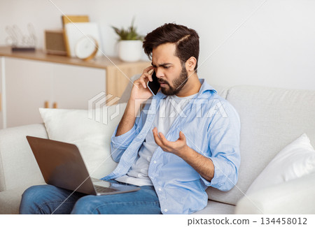 Computer Problems. Frustrated arab freelancer guy talking on cellphone and looking on laptop screen at home, calling customer support hotline, suffering bad internet connection while working online 134458012