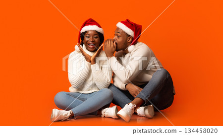 Loving Black Guy Whispering Christmas Secret To His Excited Girlfriend, Spelling To Her Ear While Having Fun Together Over Orange Background In Studio, Enjoying Celebrating Winter Holidays Together 134458020