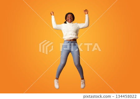 Big Win. Excited Happy African American Woman Celebrating Success While Jumping In Air, Cheerful Black Lady Raising Fists And Shouting With Joy, Having Fun Over Orange Background, Copy Space 134458022