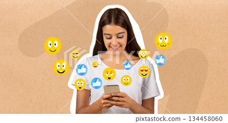 A young woman is smiling as she chats on her smartphone, surrounded by cheerful emojis. The setting conveys a sense of happiness and engagement with friends online. 134458060