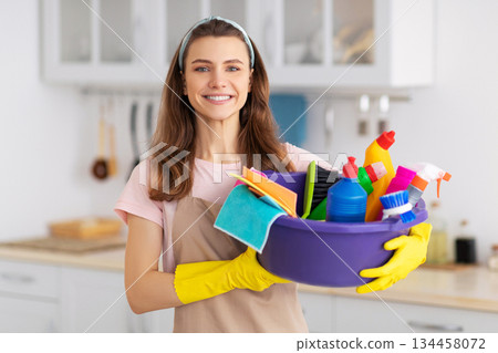 Portrait of cheerful young housewife holding cleaning supplies, smiling and looking at camera in kitchen. Professional cleaning service specialist offering help with domestic duties 134458072