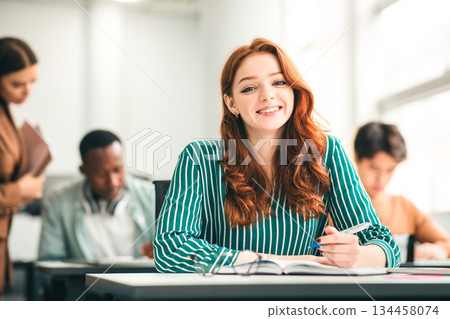 Modern Education Concept. Portrait of smiling ginger red haired female student sitting at desk in classroom at university, taking test or writing notes in her notebook, looking posing at camera Modern Education Concept. Portrait of smiling ginger red haired female student sitting at desk in classroom at university, taking test or writing notes in her notebook, looking posing at camera 134458074