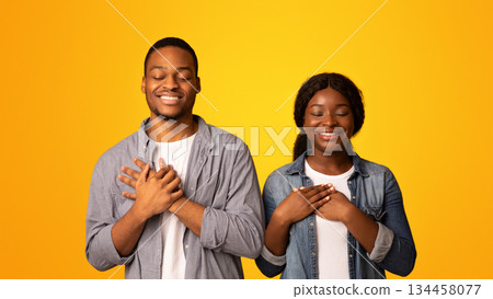 With Faith In Heart. Black couple praying with hands on chest and closed eyes, blessed african american man and woman standing isolated over yellow background, cherishing hope for best 134458077
