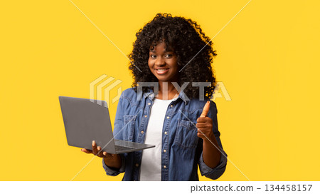 Happy african american young woman holding newest laptop and showing thumb up, yellow studio background, panorama with copy space. Cheerful black lady using notebook, shopping online or websurfing 134458157