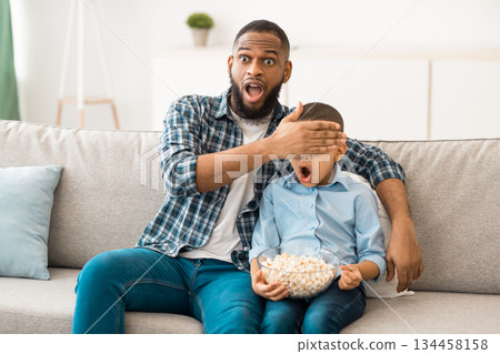 Questionable TV Content. Shocked Black Father Covering Son's Eyes Watching Television Together Sitting On Sofa At Home. Children Tv Programming Issues, Parental Control Concept Questionable TV Content. Shocked Black Father Covering Son's Eyes Watching Television Together Sitting On Sofa At Home. Children Tv Programming Issues, Parental Control Concept 134458158