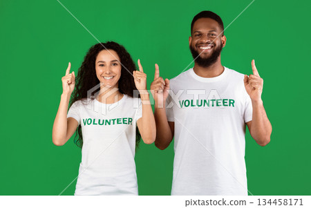 Two Multiracial Volunteers Pointing Fingers Up Advertising Something Standing Over Green Background In Studio, Smiling To Camera. Look There. Charity And Volunteering Ad Banner 134458171