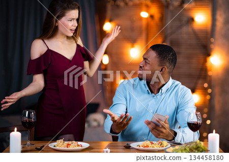 Furious Young Woman Blaming Her Black Boyfriend For Using Smartphone During Romantic Dinner. Young Interracial Couple Emotionally Arguing At Valentine's Day Date In Restaurant, Free Space 134458180