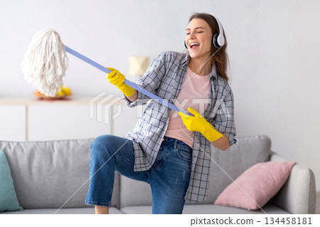 Positive young housewife wearing headphones, listening to music, playing mop like guitar while doing house cleanup. Millennial woman enjoying domestic chores, having fun while cleaning her home Positive young housewife wearing headphones, listening to music, playing mop like guitar while doing house cleanup. Millennial woman enjoying domestic chores, having fun while cleaning her home 134458181