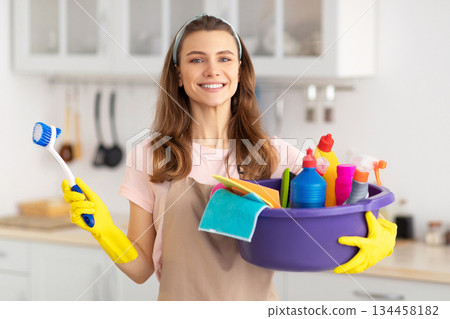 Portrait of joyful young maid with cleaning supplies smiling at camera in kitchen. Happy housewife ready to tidy her home, doing domestic duties, prepared for house work Portrait of joyful young maid with cleaning supplies smiling at camera in kitchen. Happy housewife ready to tidy her home, doing domestic duties, prepared for house work 134458182