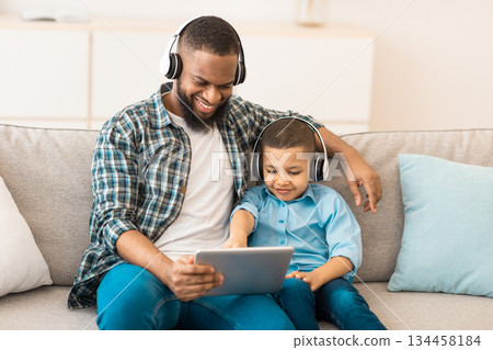 African American Dad And His Son Using Digital Tablet Wearing Headphones Watching Cartoons Online Sitting On Sofa At Home. Family Bonding Using Computer Together On Weekend 134458184