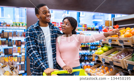 Shopping together is fun. Happy black couple choosing healthy food at fruits and veggies section of supermarket. Millennial African American family with trolley purchasing groceries at mall 134458195