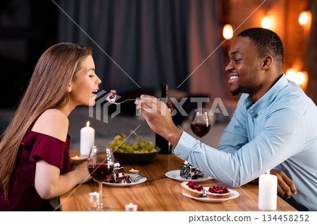 Happy Interracial Couple Having Romantic Moments At Dinner In Restaurant, Eating Desserts And Drinking Wine. Loving African American Man Feeding His White Girlfriend With Cake, Enjoying Time Together Happy Interracial Couple Having Romantic Moments At Dinner In Restaurant, Eating Desserts And Drinking Wine. Loving African American Man Feeding His White Girlfriend With Cake, Enjoying Time Together 134458252