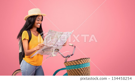 Lovely African American woman with bicycle looking at map over pink studio background. Pretty black lady in straw hat traveling by bike, plotting her tourist route 134458254