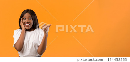 Amazed african american woman with beautiful hair pointing with thumb up at empty space, touching her face in surprised gesture and opening mouth, orange studio background, panorama 134458263