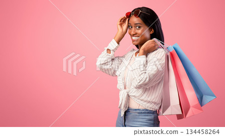 Gorgeous young woman with shopping bags smiling and looking at camera on pink studio background. Pretty black lady buying gifts for holiday, happy over big discounts 134458264