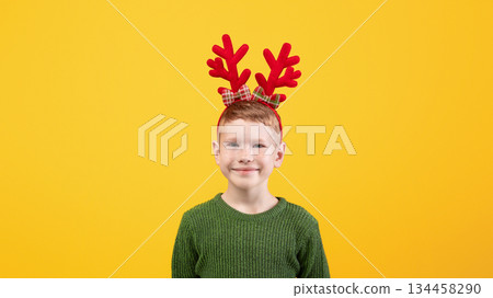 Cute kid in green winter sweater posing on yellow studio background, copy space. Happy ginger boy with deer accessory on his head waiting for winter holidays. Christmas, New Year concept 134458290