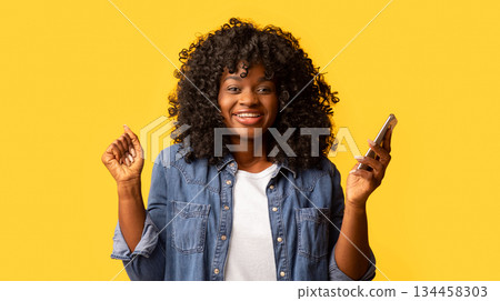 Excited black woman with smartphone raising hands up over yellow studio background, copy space. Emotional african american curly lady enjoying nice mobile application or gambling online 134458303