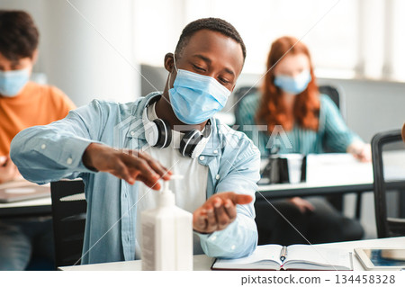 Hygiene And Protection Concept. Black student guy applying antibacterial sanitizer on hands from bottle, wearing surgical face mask, sitting in classroom. Prevent the spread of infection and germs 134458328