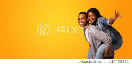 Joyful african woman piggybacking her boyfriend and showing peace gesture at camera, millennial black couple having fun together while standing isolated over yellow background, copy space 134458331