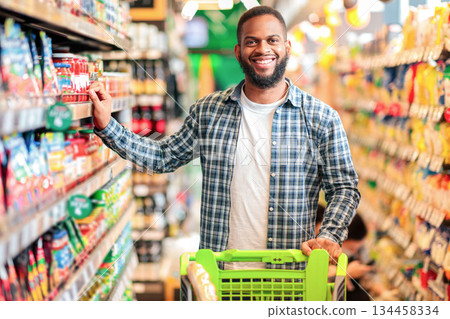 Happy African American Guy Posing In Supermarket. Cheerful Customer Choosing Food And Buying Groceries Standing Near Shelf In Local Store Indoors, Smiling To Camera. Selective Focus 134458334