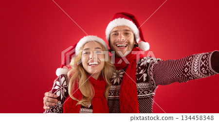 Happy young guy with his girlfriend in Christmas outfits taking selfie over red background 134458348