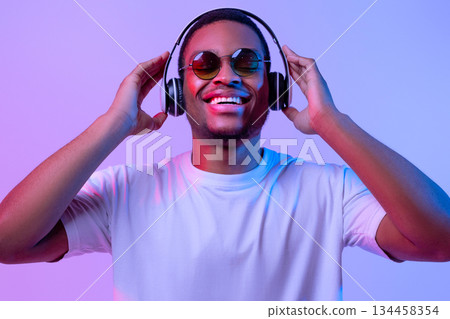 Portrait Of Cheerful African American Guy In Wireless Headphones And Sunglasses In Neon Light, Happy Handsome Black Man In White T-Shirt Standing Over Purple Studio Background, Closeup With Free Space 134458354