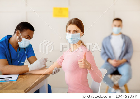 Young woman in medical mask approving of covid-19 vaccination, showing thumb up gesture at clinic. Millennial lady recommending coromavirus immunization at health centre 134458357
