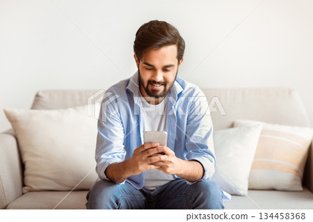 Happy arab man using smartphone at home, messaging with friends while sitting on couch in living room, young eastern guy using mobile phone, relaxing on sofa and enjoying mobile communication 134458368