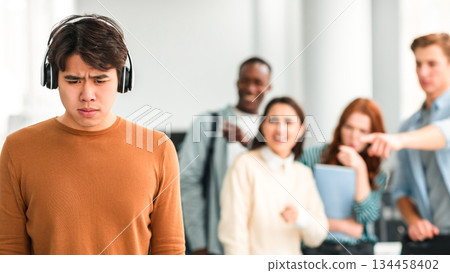Bullying Concept. Rude diverse group of people mocking and making fun of sad asian guy wearing headphones, pointing fingers at him and laughing, standing in blurred background. Prank and abuse Bullying Concept. Rude diverse group of people mocking and making fun of sad asian guy wearing headphones, pointing fingers at him and laughing, standing in blurred background. Prank and abuse 134458402