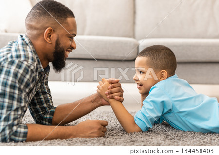 Happy African American Dad And Kid Son Arm Wrestling Competing Lying On Floor At Home. Happy Young Daddy And Little Boy Armwrestling Having Fun Together. Side View 134458403