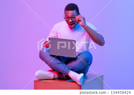 Excited Black Male In Eyeglasses Sitting On Big Cube With Laptop, Looking At Screen With Amazement, African American Student Guy Posing In Neon Light Over Purple Studio Background, Free Space Excited Black Male In Eyeglasses Sitting On Big Cube With Laptop, Looking At Screen With Amazement, African American Student Guy Posing In Neon Light Over Purple Studio Background, Free Space 134458424