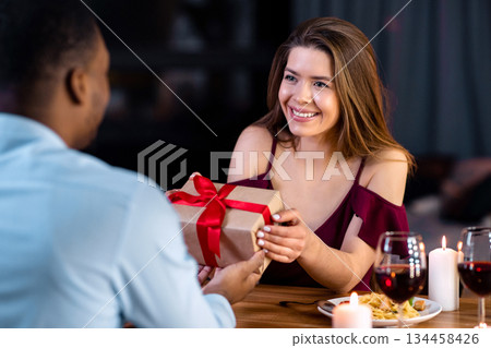 Happy Young Woman Getting Gift From Her Black Boyfriend On Romantic Dinner In Restaurant, Loving African American Man Greeting His Girfriend With International Women's Day, Selective Focus On Lady Happy Young Woman Getting Gift From Her Black Boyfriend On Romantic Dinner In Restaurant, Loving African American Man Greeting His Girfriend With International Women's Day, Selective Focus On Lady 134458426