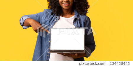 Happy beautiful black woman pointing at laptop with empty screen over yellow studio background, copy space. Cute african american young lady showing notebook with blank display, mockup 134458445