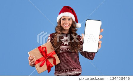 Smiling girl in Christmas outfit holding gift box and cellphone with blank screen over blue background, space for design. Panorama 134458446