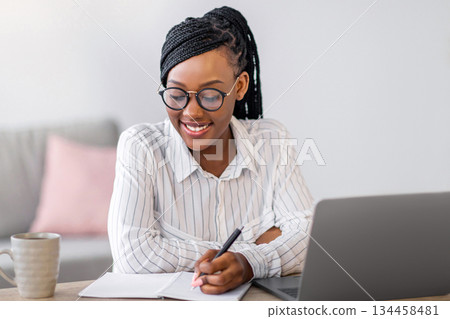 Positive black business woman with stylish glasses manager working from home during COVID-19 pandemic, using modern laptop and taking notes, sitting at workdesk in living room, copy space Positive black business woman with stylish glasses manager working from home during COVID-19 pandemic, using modern laptop and taking notes, sitting at workdesk in living room, copy space 134458481