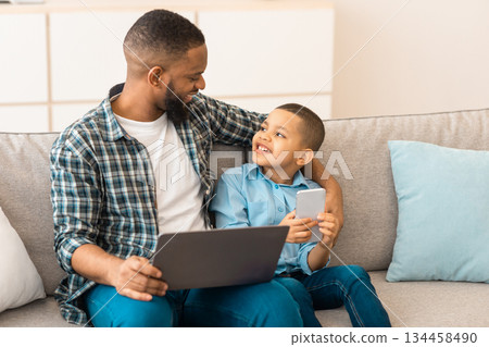 Family Gadgets. African Father And Little Son Using Laptop And Mobile Phone Browsing Internet Sitting On Sofa Together At Home. Technology Concept. Selective Focus 134458490