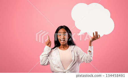 Excited black lady holding empty speech bubble and pointing her finger up on pink studio background, mockup for your design. Happy African American woman with blank word box 134458491