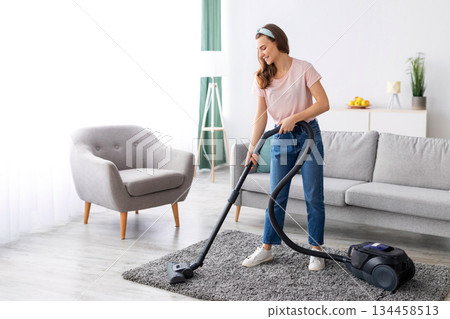 Cheerful female housekeeper using vacuum cleaner to tidy apartment, full length portrait. Positive young woman doing house chores, keeping her home clean. Domestic hygiene concept Cheerful female housekeeper using vacuum cleaner to tidy apartment, full length portrait. Positive young woman doing house chores, keeping her home clean. Domestic hygiene concept 134458513