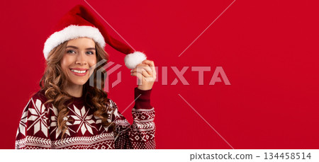 Portrait of pretty smiling woman in Santa hat and Christmas sweater over red studio background, panorama. Lovely young lady ready for Xmas, New Year celebration and winter holidays 134458514