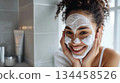 A joyful young woman with a skincare mask on her face is holding her cheeks while in a modern bathroom. A joyful young woman with a skincare mask on her face is holding her cheeks while in a modern bathroom. 134458526