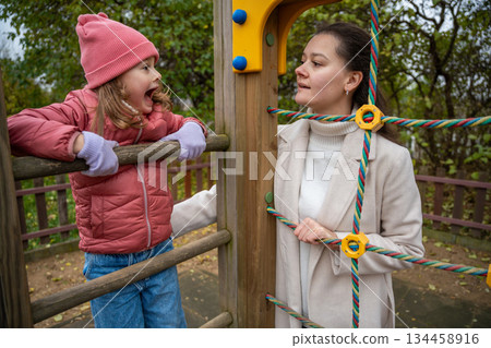 Mother and daughter talk and laugh while playing on a rope structure at the playground. Communication, joy and family time outdoors. 134458916