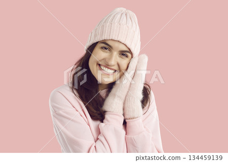 Smiling brunette woman in knitted hat and mittens, pink sweatshirt on pink background. Smiling brunette woman in knitted hat and mittens, pink sweatshirt on pink background. 134459139