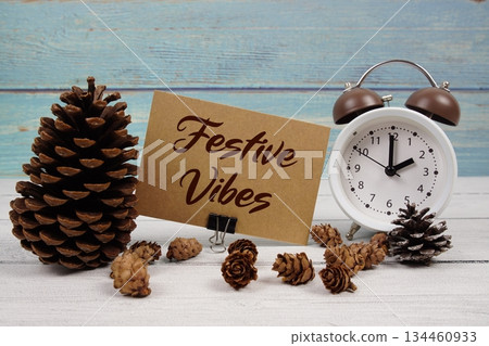 Festive Vibes text written on paper card with alarm clock and pine cones decoration on wooden background Festive Vibes text written on paper card with alarm clock and pine cones decoration on wooden background 134460933