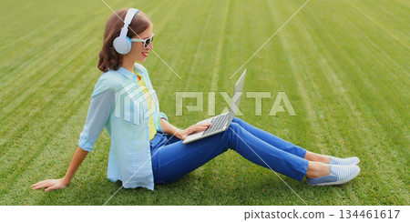 Happy young woman with laptop listening to music in headphones in summer park 134461617